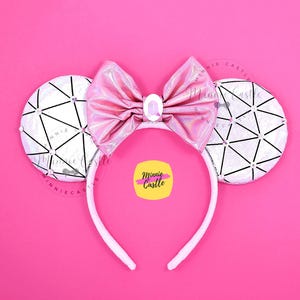 Könnte beinhalten: Ein weißes Stirnband mit angebrachten Mauseohren und einer rosa Schleife. Die Ohren sind weiß mit einem geometrischen schwarzen Design und Strasssteinen. Die Schleife ist rosa mit einem kleinen, ovalen Schmuck in der Mitte. Der Hintergrund ist rosa.