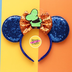 Puede incluir: Orejas de Minnie Mouse de lentejuelas azules y naranjas con un lazo de sombrero de Mickey Mouse verde y blanco. Las orejas están en una diadema azul. La diadema tiene el texto "Minnie Castle".