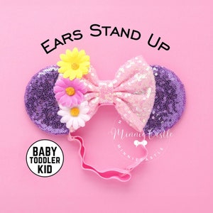 May include: Purple and pink sequined Minnie Mouse ears with a pink bow and a flower embellishment. The ears are attached to a pink headband. The text "EARS STAND UP" is written above the ears. The text "BABY TODDLER KID" is written in a circle below the ears.