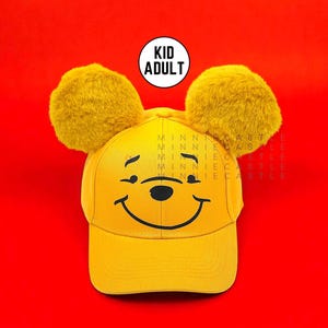 May include: Yellow baseball cap with a cartoon bear face and two fluffy yellow ears. The cap has a curved brim and a smiling face design. The text "KID ADULT" is in a white circle on the top. The background is red.