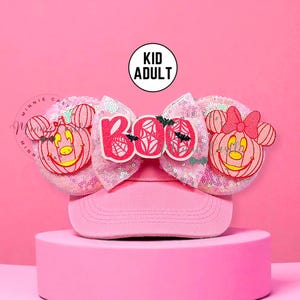 May include: Pink baseball cap with sequined Minnie Mouse ears featuring jack-o'-lantern faces. A pink bow with the word "Boo" and spiderweb and bat accents sits atop the cap. The text "Kid Adult" is in a white circle.
