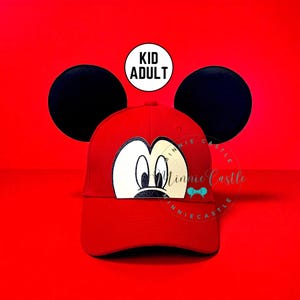 May include: A red baseball cap featuring a Mickey Mouse face design. The cap has a curved brim and a black mouse ear on each side. The text "KID ADULT" is in a white circle. The background is a solid red.