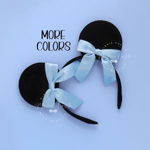 May include: Black mouse ear headband with a light blue satin bow. The text "MORE COLORS" is displayed in black. The headband is on a light blue background.