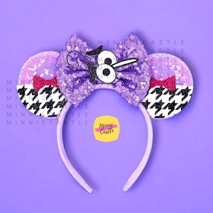 May include: A headband featuring two circular ears covered in purple sequins and a black and white houndstooth pattern. A large central purple sequin bow displays a white cartoon eye graphic. Small red bows accent the base of each ear.