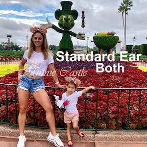 Puede incluir: Dos personas con camisetas blancas a juego con un gr&aacute;fico de Minnie Mouse rosa y orejas de Minnie Mouse de oro rosa. La mujer lleva pantalones cortos de mezclilla y la ni&ntilde;a lleva pantalones cortos rosas. Est&aacute;n de pie frente a una escultura de Mickey Mouse de topiario verde y un macizo de flores rojas.