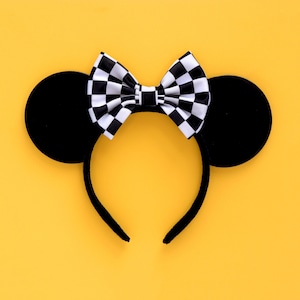 May include: Black mouse ears headband with a checkered bow. The headband is on a yellow background.