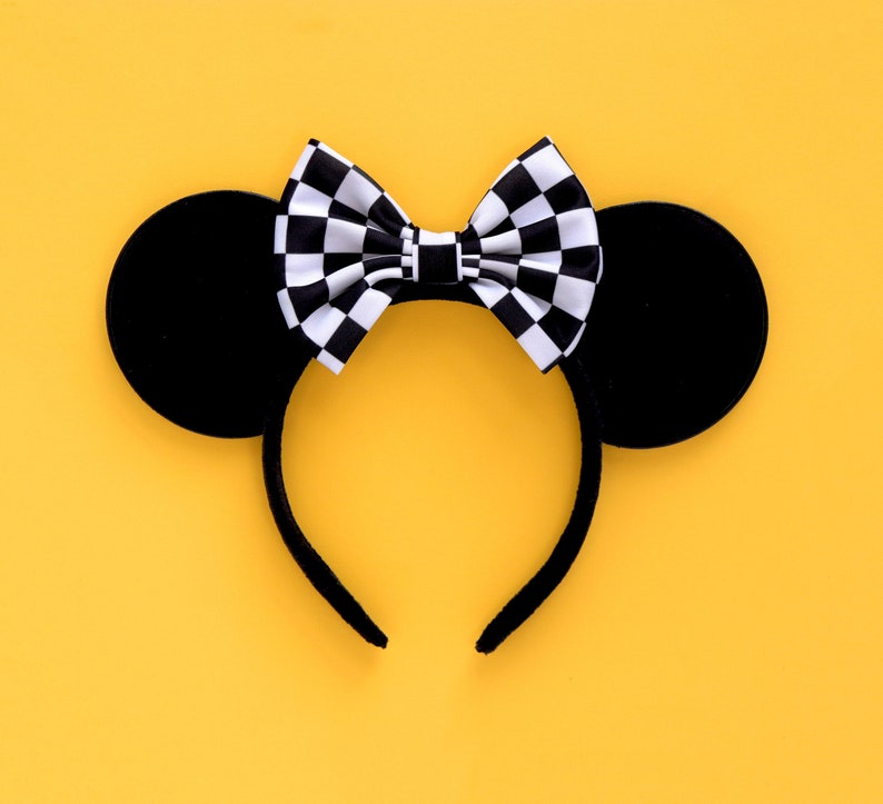May include: Black mouse ears headband with a checkered bow. The headband is on a yellow background.