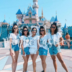 May include: Five friends wearing white t-shirts with different text and blue denim shorts pose in front of a blue and white castle. The bride-to-be is wearing a white veil and her shirt says "happily ever after" with a Mickey Mouse head. The other shirts say "I'm so ready to be a wifey" and "Let's get this castle party started".