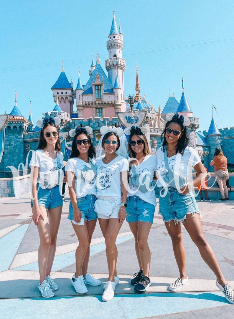 May include: Five friends wearing white t-shirts with different text and blue denim shorts pose in front of a blue and white castle. The bride-to-be is wearing a white veil and her shirt says "happily ever after" with a Mickey Mouse head. The other shirts say "I'm so ready to be a wifey" and "Let's get this castle party started".