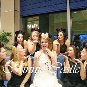 K&ouml;nnte beinhalten: Eine Gruppe von Frauen in schwarzen Kleidern und Minnie Mouse Ohren posiert f&uuml;r ein Foto. Die Frau in der Mitte tr&auml;gt ein wei&szlig;es Kleid und eine Tiara. Der Text "Minnie Castle" ist im Vordergrund geschrieben.