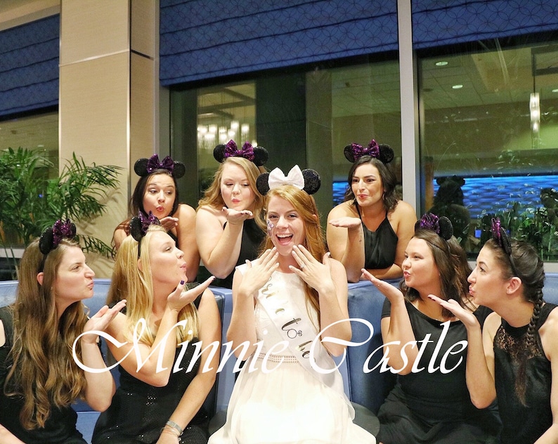 K&ouml;nnte beinhalten: Eine Gruppe von Frauen in schwarzen Kleidern und Minnie Mouse Ohren posiert f&uuml;r ein Foto. Die Frau in der Mitte tr&auml;gt ein wei&szlig;es Kleid und eine Tiara. Der Text "Minnie Castle" ist im Vordergrund geschrieben.