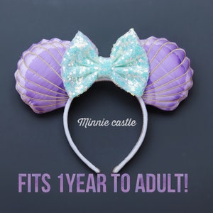 May include: A headband with seashell-shaped ears and a sequined bow. The ears are purple with gold trim, and the bow is light blue. The text "Minnie castle" is printed on the headband. The text "FITS 1YEAR TO ADULT!" is printed below the headband.
