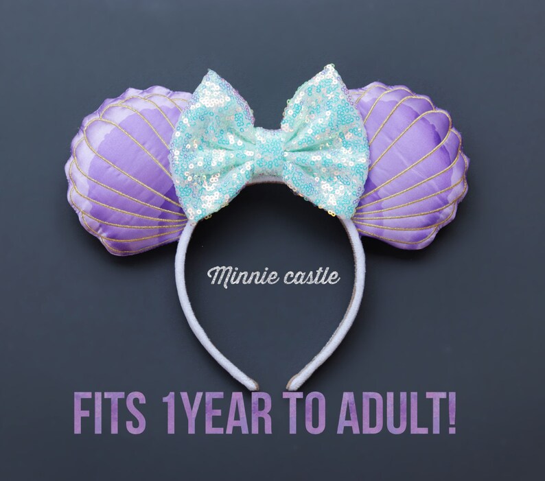 May include: A headband with seashell-shaped ears and a sequined bow. The ears are purple with gold trim, and the bow is light blue. The text "Minnie castle" is printed on the headband. The text "FITS 1YEAR TO ADULT!" is printed below the headband.