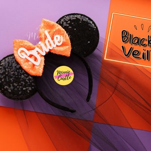 May include: Black and orange Minnie Mouse ears with a white "Bride" bow and a black tulle veil. The ears are decorated with black sequins. The veil is labeled "Black Veil".