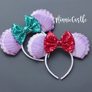 May include: Two headbands with seashell designs. One has a teal sequin bow, the other a red sequin bow. The seashell ears are lavender with gold stitching. The headbands are white and the background is gray. The text "MinnieCastle" is in the upper right corner.