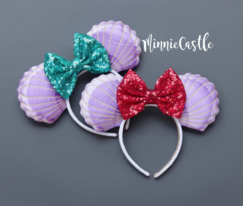 May include: Two headbands with seashell designs. One has a teal sequin bow, the other a red sequin bow. The seashell ears are lavender with gold stitching. The headbands are white and the background is gray. The text "MinnieCastle" is in the upper right corner.