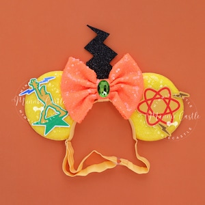 Op de afbeelding: Gele Minnie Mouse oren met een zwarte bliksem, een oranje strik met een groene edelsteen en een groene, gele en rode bliksem en atoom afbeelding.