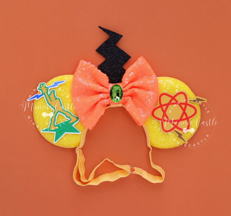 Op de afbeelding: Gele Minnie Mouse oren met een zwarte bliksem, een oranje strik met een groene edelsteen en een groene, gele en rode bliksem en atoom afbeelding.