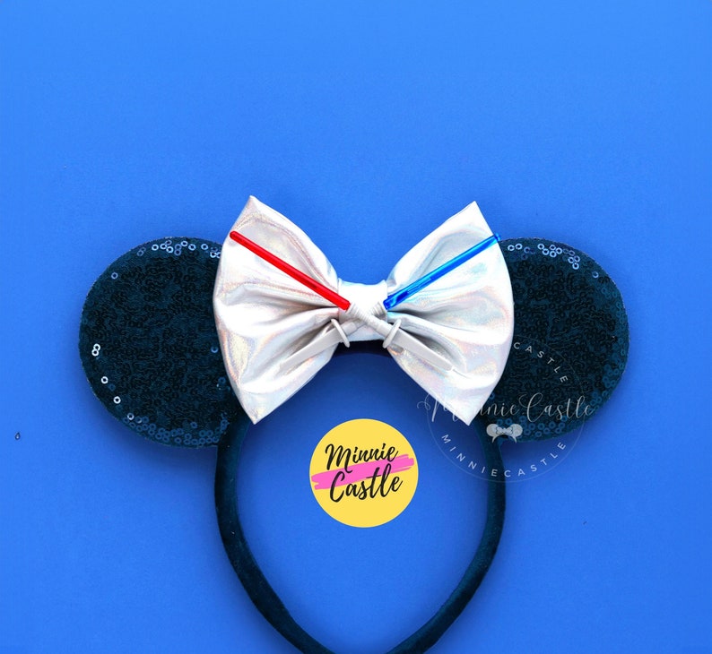 Puede incluir: Un par de orejas de Minnie Mouse de color azul oscuro brillante con un lazo plateado y dos sables de luz que se iluminan. El lazo tiene un centro blanco y los sables de luz son rojo y azul. Las orejas est&aacute;n en una diadema negra. La diadema tiene un c&iacute;rculo amarillo con el texto "Minnie Castle" en &eacute;l.
