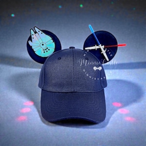 May include: Navy blue baseball cap with attached mouse ears featuring a Millennium Falcon design and lightsaber. The cap has the text "Minnie Castle" on the front.