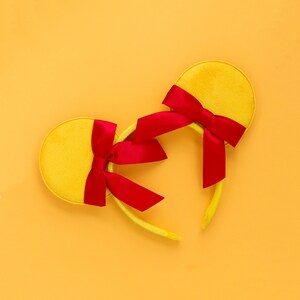 May include: A yellow mouse ear headband with red satin bows. The headband has two circular yellow ears and a matching yellow band. The red bows are tied at the base of each ear, creating a playful and festive accessory.
