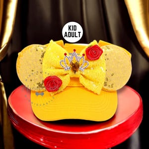 May include: Yellow baseball cap with sequined mouse ears, a bow, and a crown. The bow is adorned with a jeweled crown and red roses. The text "KID ADULT" is in a white circle.