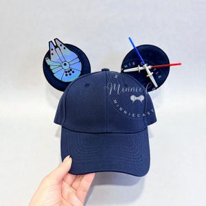 May include: Navy blue baseball cap with attached black Mickey Mouse ears. One ear features a Millennium Falcon design, the other a lightsaber. The cap has the text "Minnie Castle" in a cursive font.