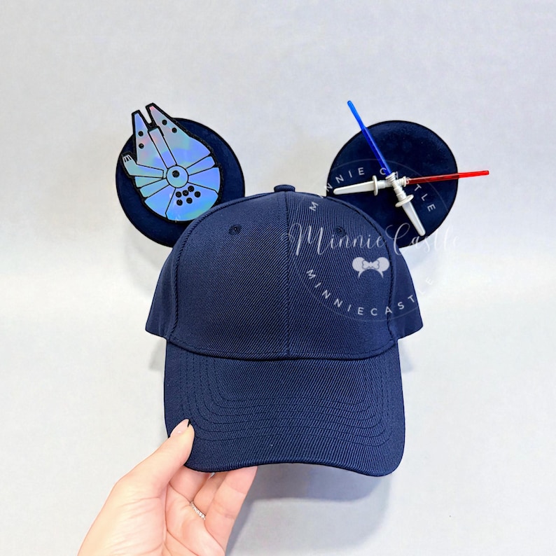 May include: Navy blue baseball cap with attached black Mickey Mouse ears. One ear features a Millennium Falcon design, the other a lightsaber. The cap has the text "Minnie Castle" in a cursive font.