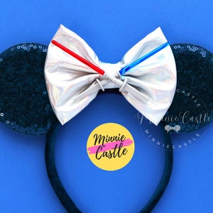 Puede incluir: Un par de orejas de Minnie Mouse de lentejuelas negras con un lazo plateado iridiscente y dos sables de luz rojos y azules. El lazo est&aacute; sujeto a la diadema con un clip plateado. La diadema es negra y tiene el texto "Minnie Castle" en un c&iacute;rculo amarillo con un contorno rosa.
