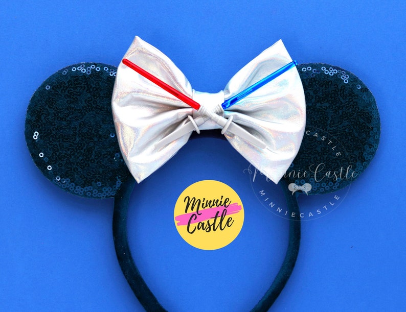 Puede incluir: Un par de orejas de Minnie Mouse de lentejuelas negras con un lazo plateado iridiscente y dos sables de luz rojos y azules. El lazo est&aacute; sujeto a la diadema con un clip plateado. La diadema es negra y tiene el texto "Minnie Castle" en un c&iacute;rculo amarillo con un contorno rosa.