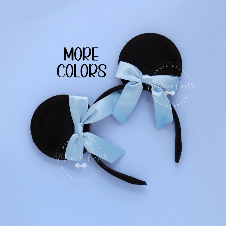 May include: Black mouse ear headband with a light blue satin bow. The text "MORE COLORS" is displayed in black. The headband is on a light blue background.