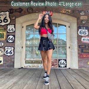 May include: A woman wearing a red crop top, black skirt, and checkered fanny pack. She is wearing a Minnie Mouse ear headband with a checkered bow and a black and white checkered pattern. The background includes Route 66 signs and the text "Customer Review Photo: 4" Large ear."