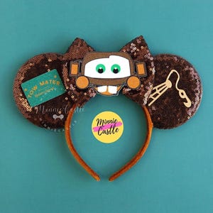 May include: Brown sequined mouse ears with a Mater character design. The ears have a brown bow and a brown headband. A green tag with the text "Tow Mater" is on one ear. A yellow sticker with the text "Minnie Castle" is on the headband.