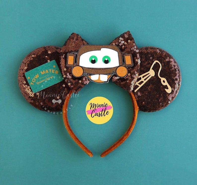 May include: Brown sequined mouse ears with a Mater character design. The ears have a brown bow and a brown headband. A green tag with the text "Tow Mater" is on one ear. A yellow sticker with the text "Minnie Castle" is on the headband.
