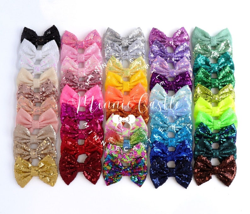 Puede incluir: Una colecci&oacute;n de 24 lazos para el pelo con lentejuelas en varios colores, incluyendo negro, blanco, rosa, rojo, naranja, amarillo, verde, azul, morado y marr&oacute;n. Los lazos est&aacute;n dispuestos en una fila, con los colores pasando de oscuro a claro.