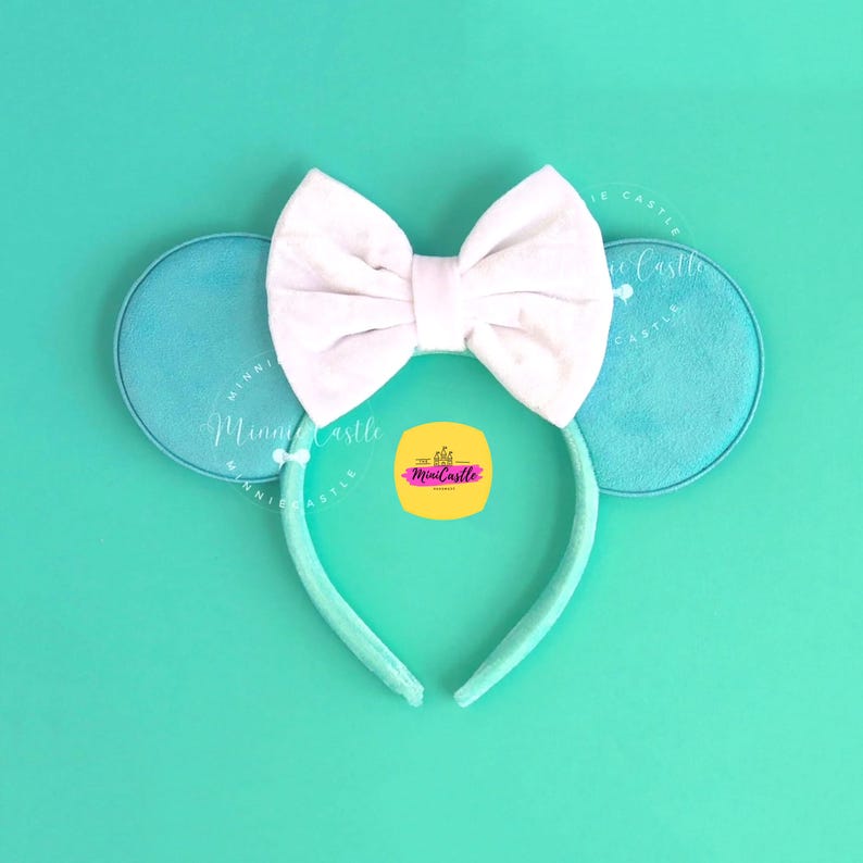 May include: A headband featuring two round, light blue ears and a large, centered white fabric bow. The headband band is covered in matching light blue fabric. A yellow square logo with the text MiniCastle is visible in the center.