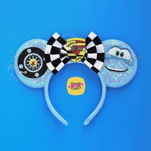 May include: Blue headband with sequined ears featuring a race car wheel and a smiling cloud. A black and white checkered bow with a car emblem is centered on the headband. The background is a solid blue.