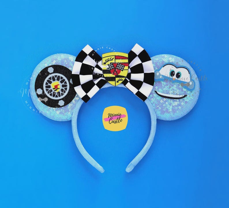 May include: Blue headband with sequined ears featuring a race car wheel and a smiling cloud. A black and white checkered bow with a car emblem is centered on the headband. The background is a solid blue.