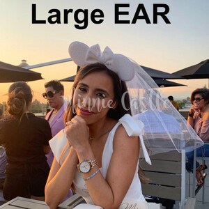 May include: A white bridal veil with a white headband featuring large sparkly Minnie Mouse ears. The headband has a white bow. The veil is draped over the woman's head and shoulders. The woman is wearing a white sleeveless dress with the word "Mrs." written on it.
