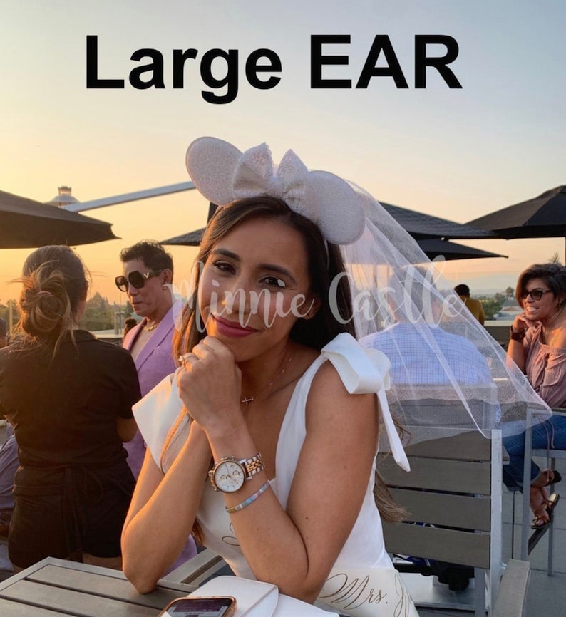 May include: A white bridal veil with a white headband featuring large sparkly Minnie Mouse ears. The headband has a white bow. The veil is draped over the woman's head and shoulders. The woman is wearing a white sleeveless dress with the word "Mrs." written on it.