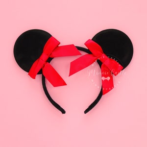May include: Black mouse ear headband with red satin bows. The ears are round and attached to a black headband. The bows are centered on each ear. The background is a solid pink colour.