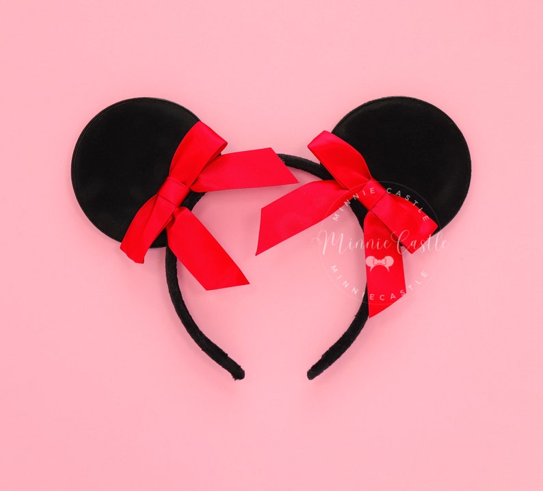 May include: Black mouse ear headband with red satin bows. The ears are round and attached to a black headband. The bows are centered on each ear. The background is a solid pink colour.