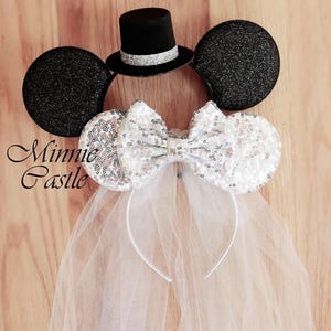 May include: Black Minnie Mouse ears with a white tulle veil and a silver sequined bow. A black top hat with a silver band sits on top of the ears. The text "Minnie Castle" is visible in the image.