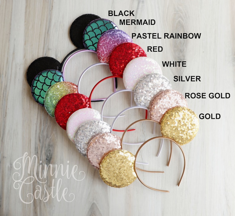 May include: A variety of Minnie Mouse ears with different colored sequins. The colors include black, mermaid, pastel rainbow, red, white, silver, rose gold, and gold. The ears are arranged in a circle on a wooden surface. The text "Minnie Castle" is written in cursive at the bottom of the image.