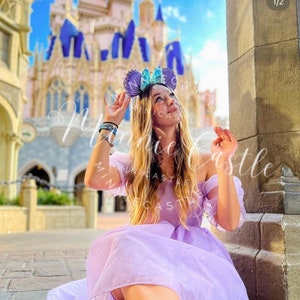 May include: A person wearing a lavender off-the-shoulder dress and a headband with purple and blue ears, posing in front of a castle. The image has the text "Minnie Castle" in a cursive font.