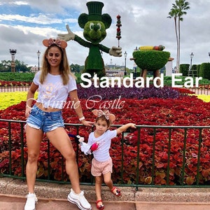Puede incluir: Una mujer y una ni&ntilde;a peque&ntilde;a con camisas blancas a juego con un dise&ntilde;o de Mickey Mouse y orejas de Minnie Mouse de oro rosa. Est&aacute;n de pie frente a una escultura de Mickey Mouse topiaria y un jard&iacute;n de flores. El texto "Standard Ear Minnie Castle" es visible en la imagen.
