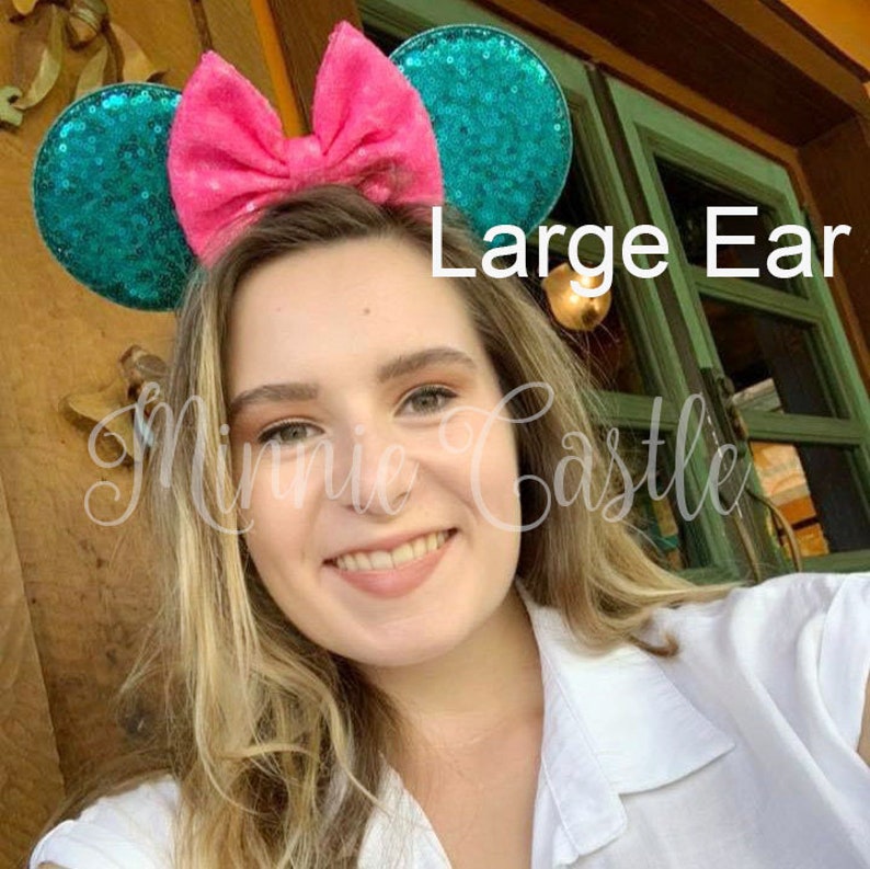Puede incluir: Una mujer lleva un par de orejas de Minnie Mouse de color turquesa y rosa con lentejuelas. Las orejas son grandes y tienen un lazo rosa en el centro. El texto "Large Ear" es visible en la imagen.