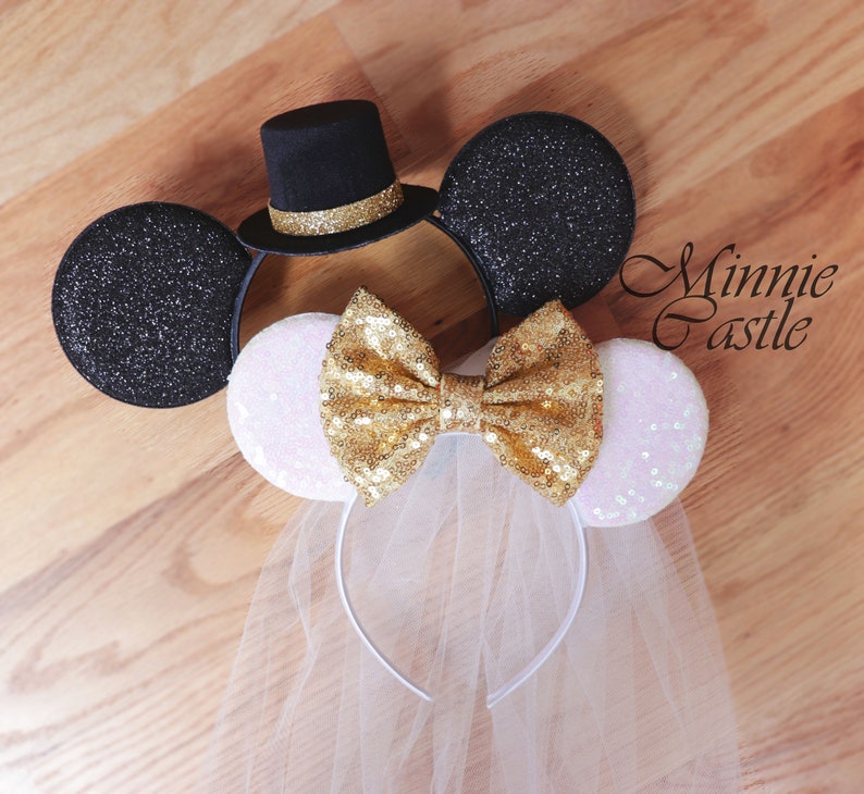 K&ouml;nnte beinhalten: Ein Paar schwarze und wei&szlig;e Minnie Mouse Ohren mit einer goldenen Schleife und einem wei&szlig;en Schleier. Die Ohren sind auf einem Haarreif. Das schwarze Ohr hat einen schwarzen Zylinder mit einem goldenen Band.