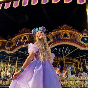 May include: A woman wearing a light purple dress with off-the-shoulder sleeves and a matching headband with mouse ears. The background features a carousel and a decorative canopy.