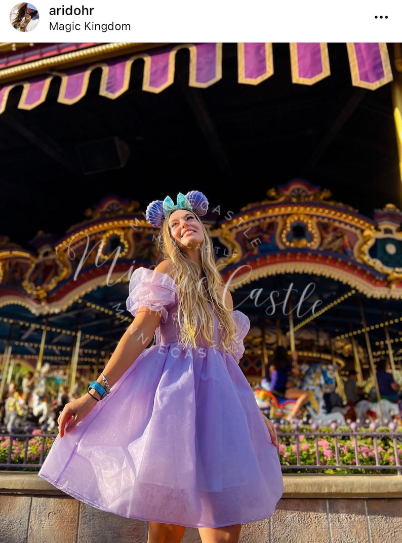 May include: A woman wearing a light purple dress with off-the-shoulder sleeves and a matching headband with mouse ears. The background features a carousel and a decorative canopy.